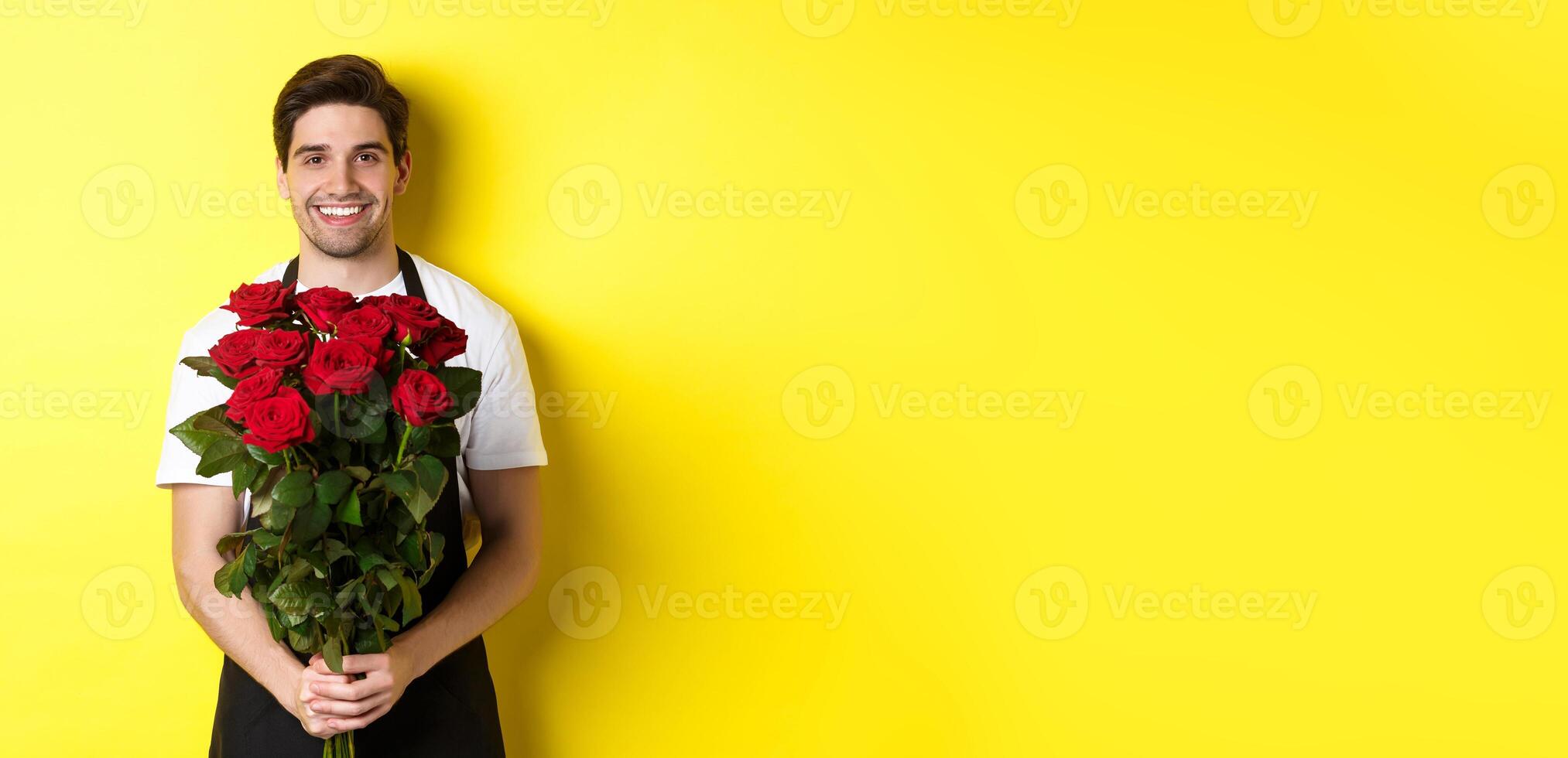 Smiling florist in black apron holding flowers, selling bouquet of roses, standing over yellow background photo