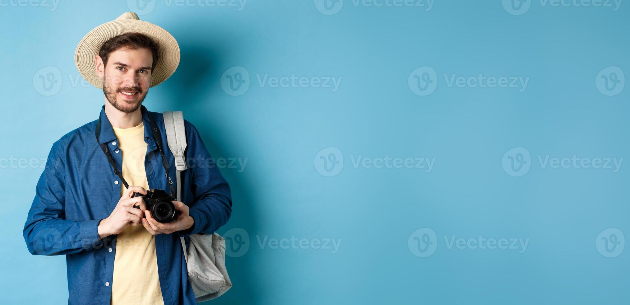 Handsome guy going on summer vacation, backpacking on holiday. Tourist with straw hat and camera smiling happy, standing on blue background photo