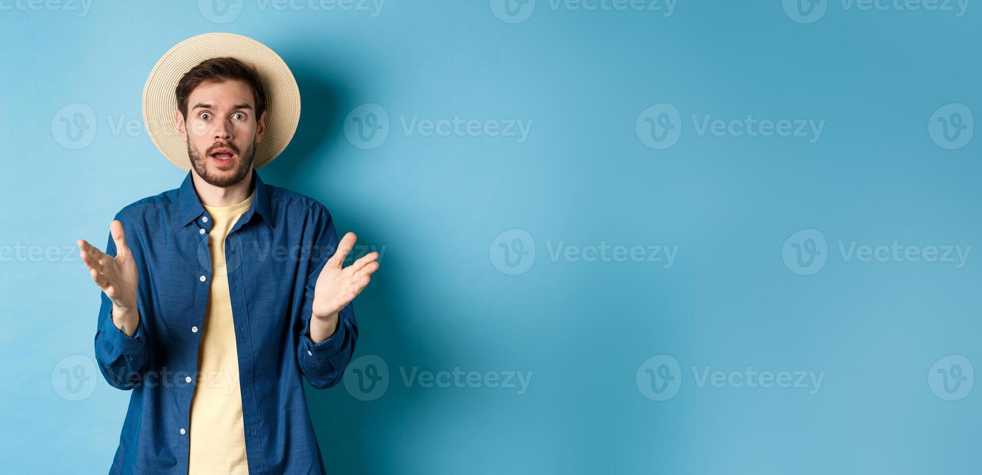 Shocked male tourist on vacation raising hands up and looking at camera startled, react to big news with disbelief, standing on blue background in straw summer hat and shirt photo