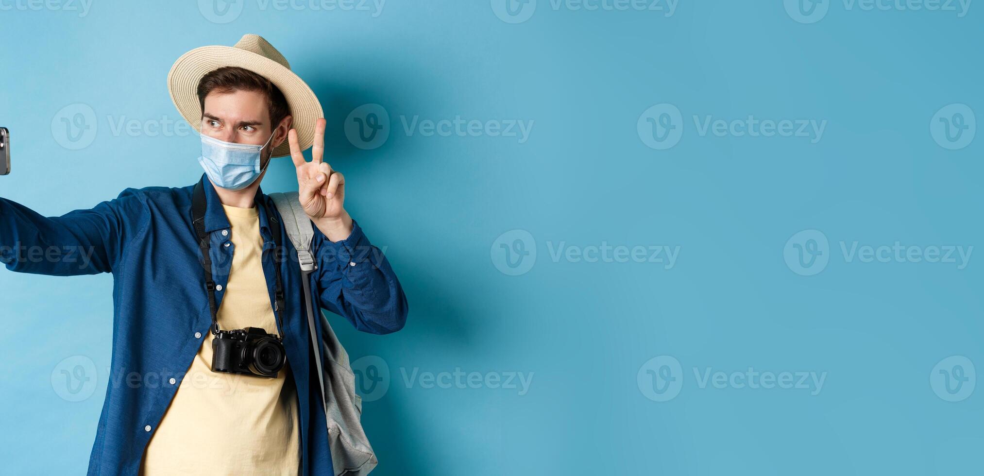 Covid-19, pandemic and travel concept. Happy and positive guy in tourist hat taking selfie and showing peace sign, posing near sightseeing, blue background photo