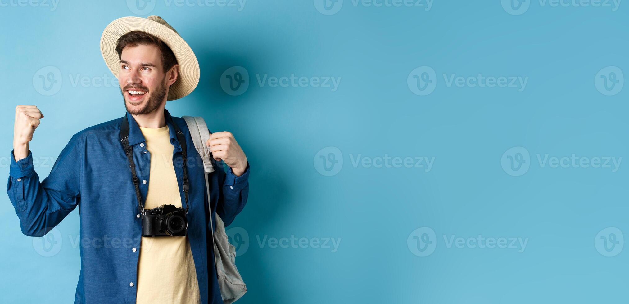 Cheerful tourist rejoicing on vacation, celebrating summer holidays, showing fist pump gesture and saying yes with satisfied face, looking aside, blue background photo
