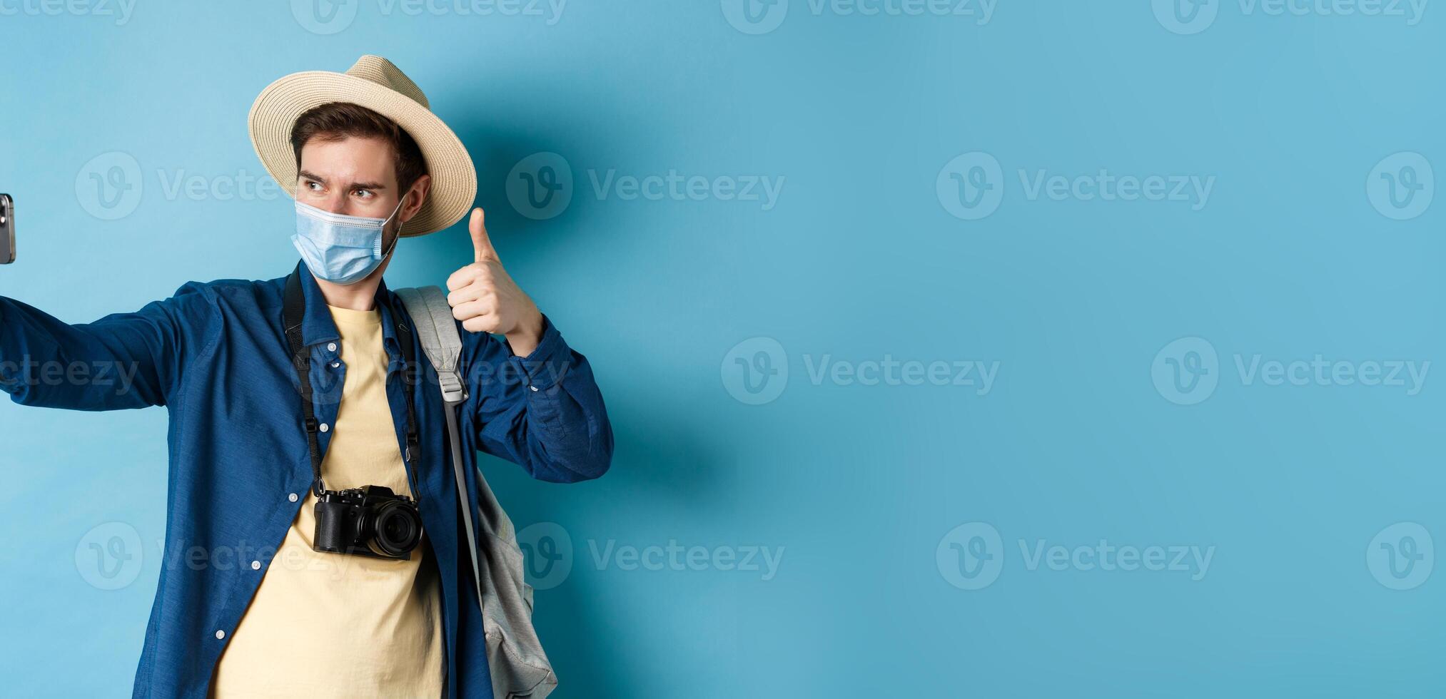 Covid-19, pandemic and travel concept. Happy male tourist taking pictures on vacation, make selfie with thumb up, recommending hotel, standing on blue background photo
