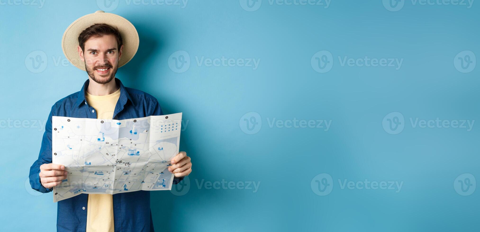 Happy and positive guy on vacation, looking at camera and holding map, smiling excited, going on summer travel, standing on blue background photo