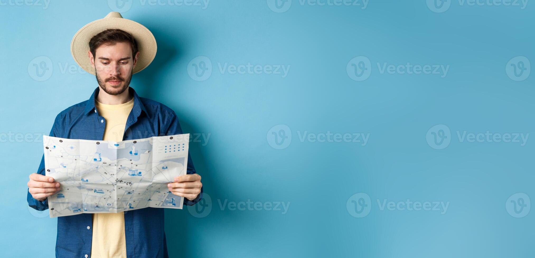 Handsome tourist with summer hat reading map, looking for sightseeing on vacation, standing on blue background photo