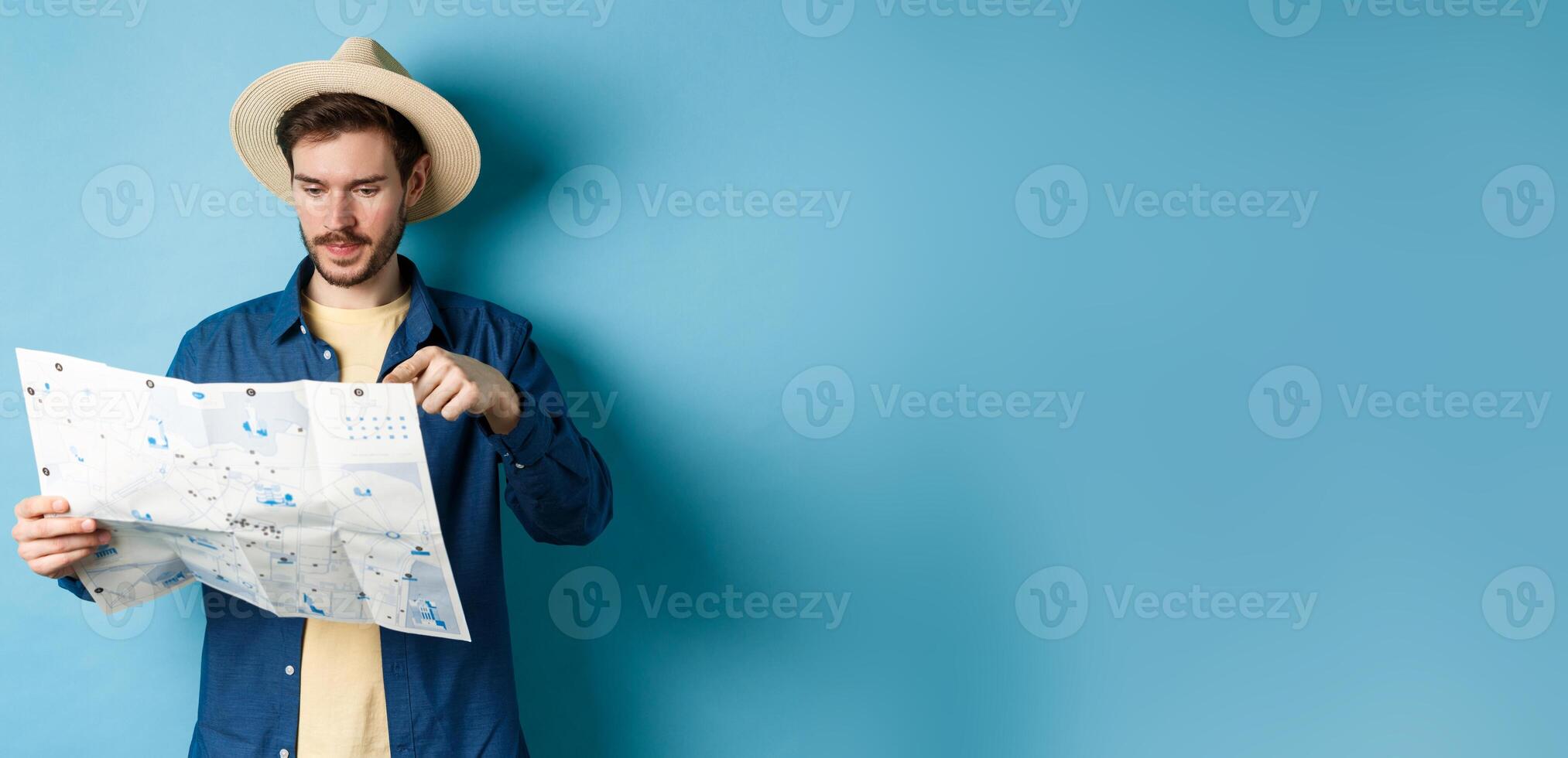 Handsome guy choosing place to go, pointing at sighseeing map on vacation, travelling on summer holidays, wearing straw hat and shirt, blue background photo