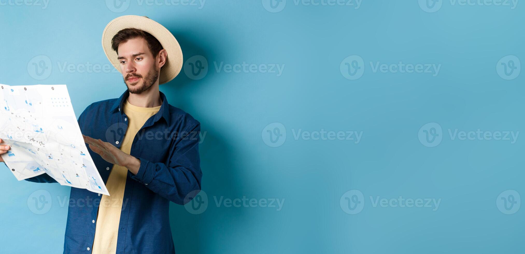 Image of handsome caucasian man looking at travel map, studying route during summer vacation, standing in straw hat on blue background photo