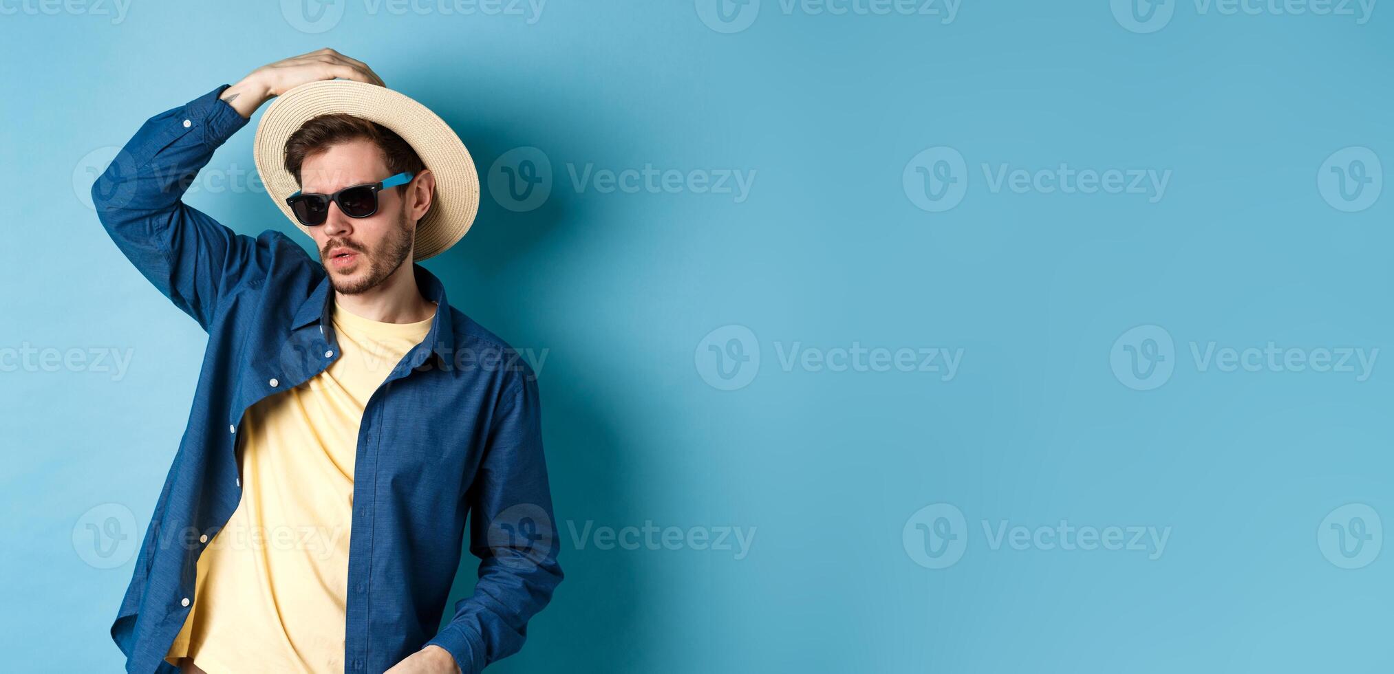 Cool guy having fun on vacation, wearing straw hat and sunglasses, looking aside sassy, standing on blue background photo