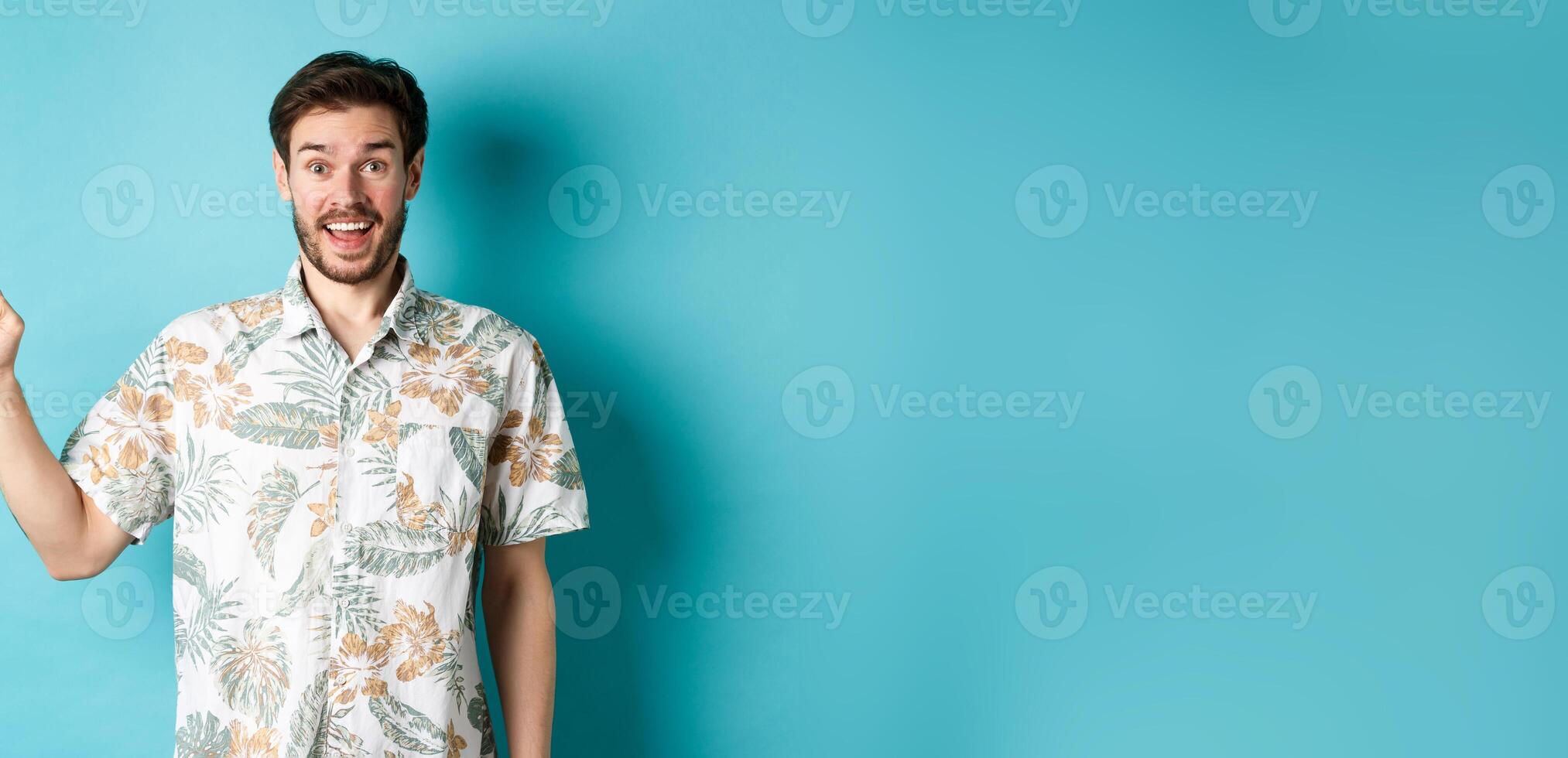 Surprised happy tourist pointing finger left at empty space, showing cool promo, standing in hawaiian shirt on blue background photo
