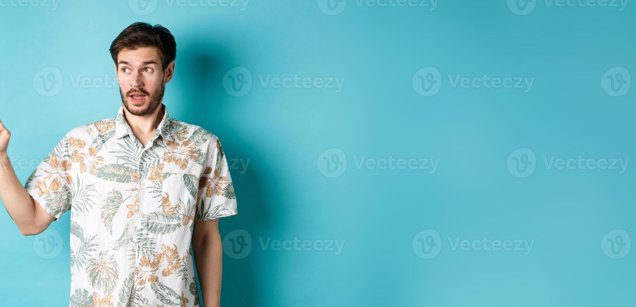 Intrigued man in hawaiian shirt, looking and pointing left at something interesting, blue background. Concept of tourism and vacation photo