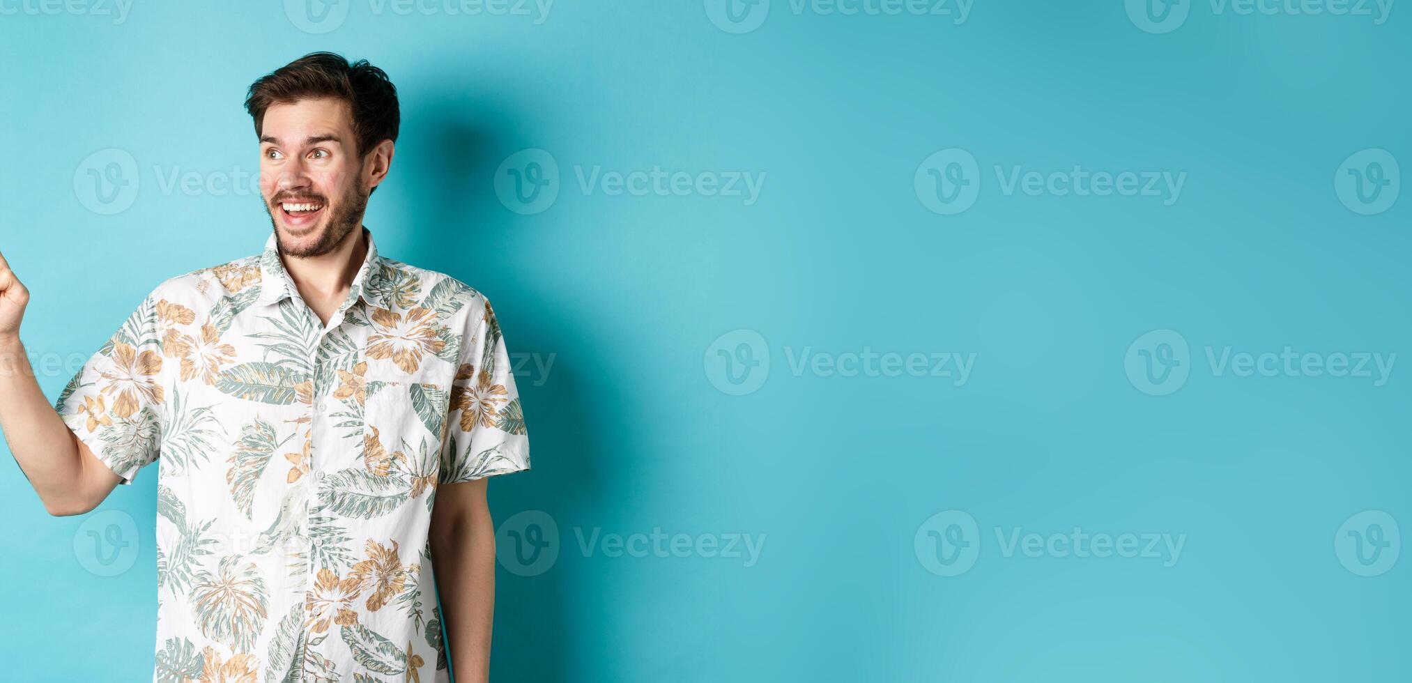 Excited smiling tourist pointing and looking aside at empty space, checking out promo offer, standing in hawaiian shirt on blue background photo