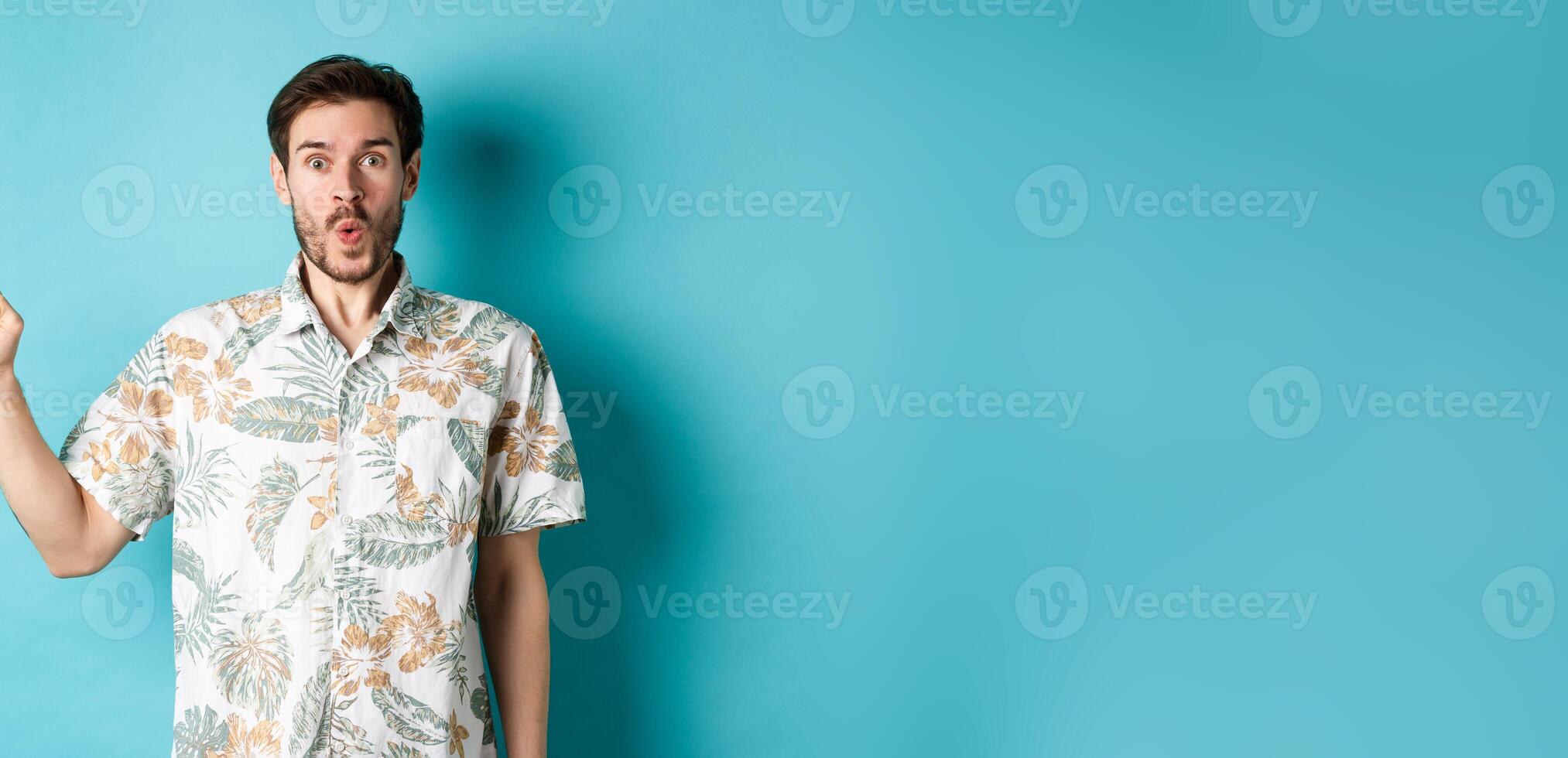 Surprised and amazed tourist in hawaiian shirt pointing finger left, showing promo and staring at camera, standing on blue background photo