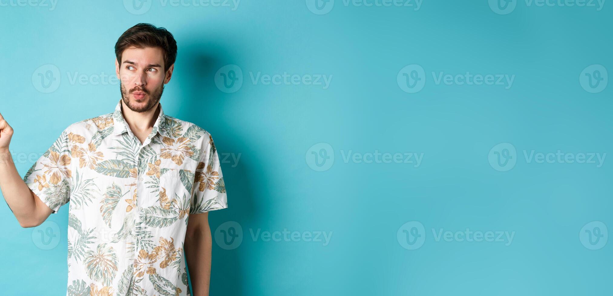 Handsome guy wearing summer shirt on vacation, pointing and looking aside at empty space, making amazed face, standing on blue background photo