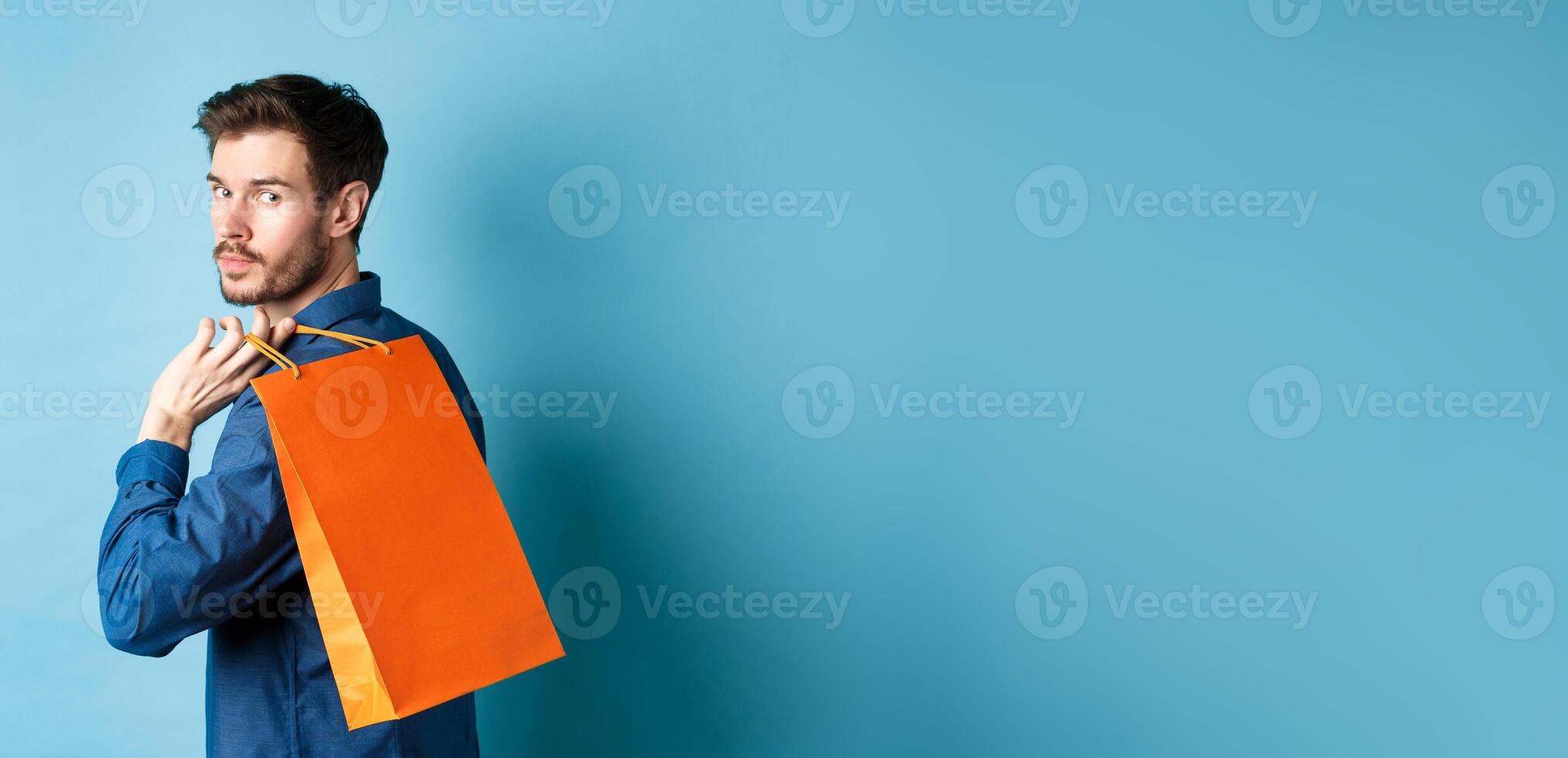 Rear view of happy man holding shopping back on shoulder and looking behind, standing on blue background photo