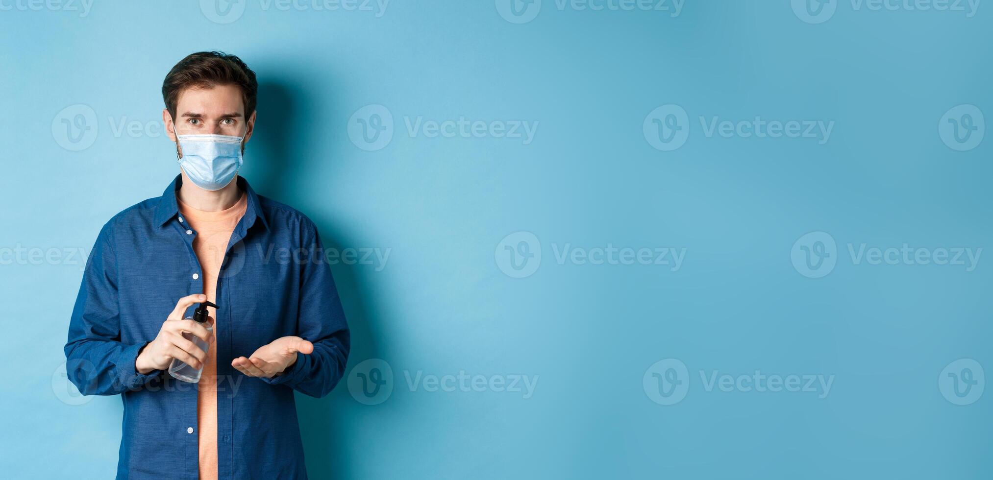 Covid-19, health and pandemic concept. Young guy in medical mask cleaning hands with sanitizer, apply antiseptic on palm, standing on blue background photo