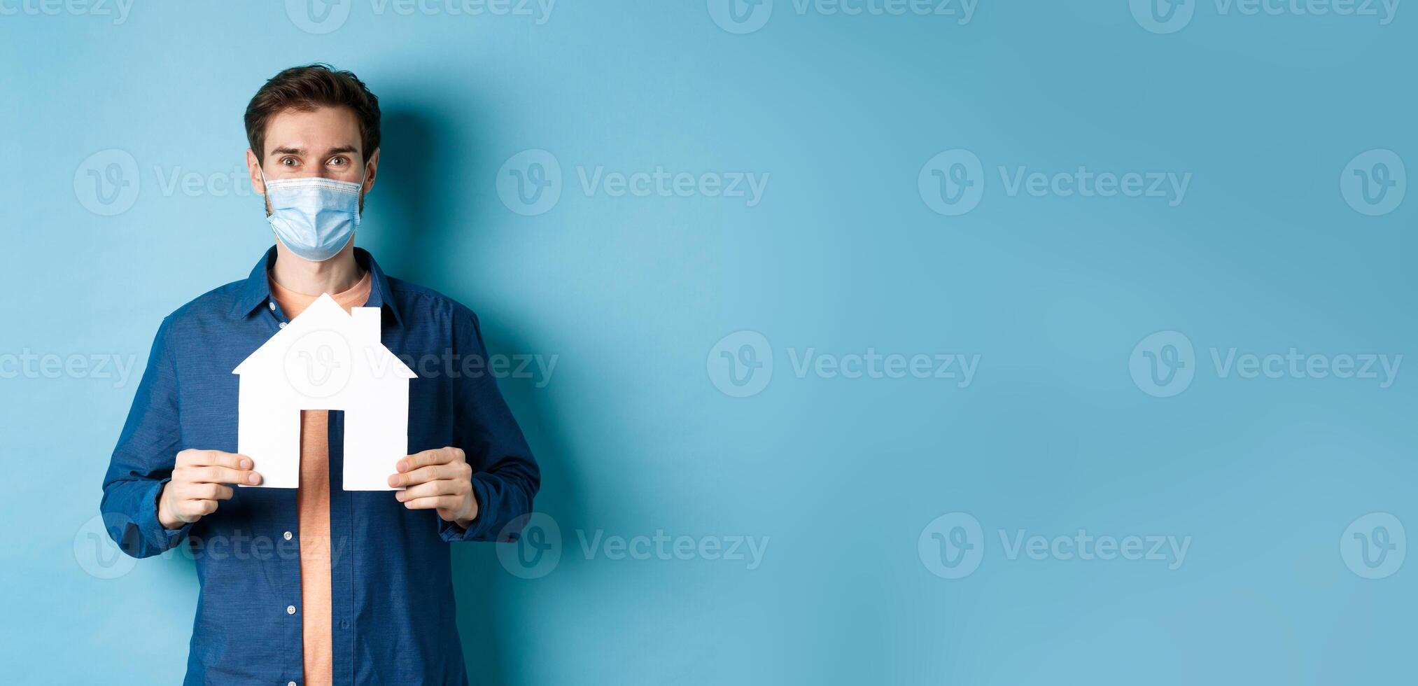 Real estate and quarantine concept. Happy young man in medical mask showing house cutout, buying property, standing on blue background photo