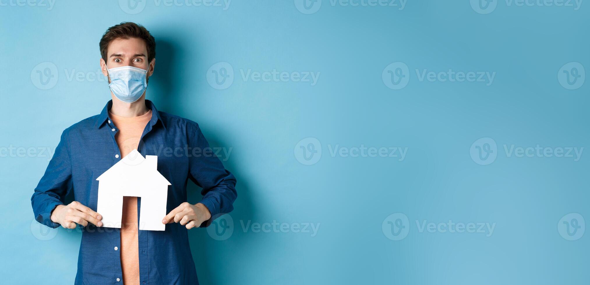 Real estate and covid concept. Happy young man in face mask demonstrate paper house cutout, looking surprised at camera, blue background photo