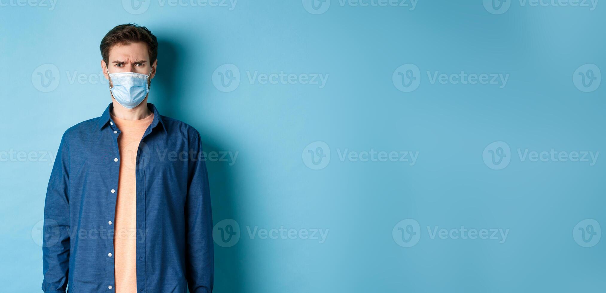 Covid-19 and healthcare concept. Disappointed guy in medical mask frowning, looking confused, standing on blue background photo
