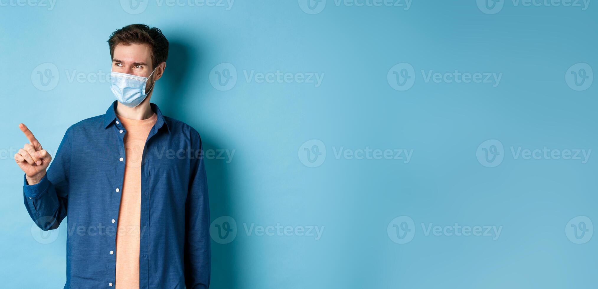 Handsome young man wearing medical mask from coronavirus, pointing finger left and looking satisfied, smiling at empty space, blue background photo