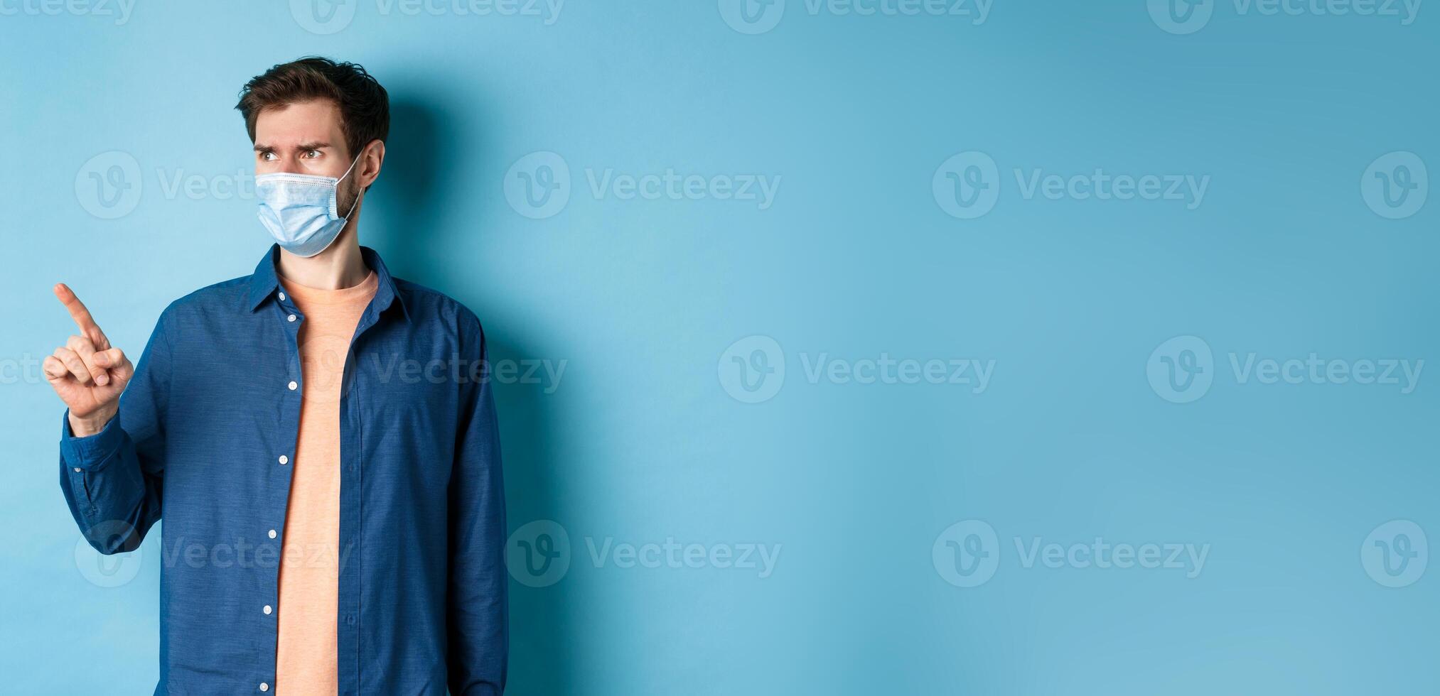 Covid-19 and healthcare concept. Confused and doubtful man in face mask frowning, pointing and looking at empty space, standing on blue background photo