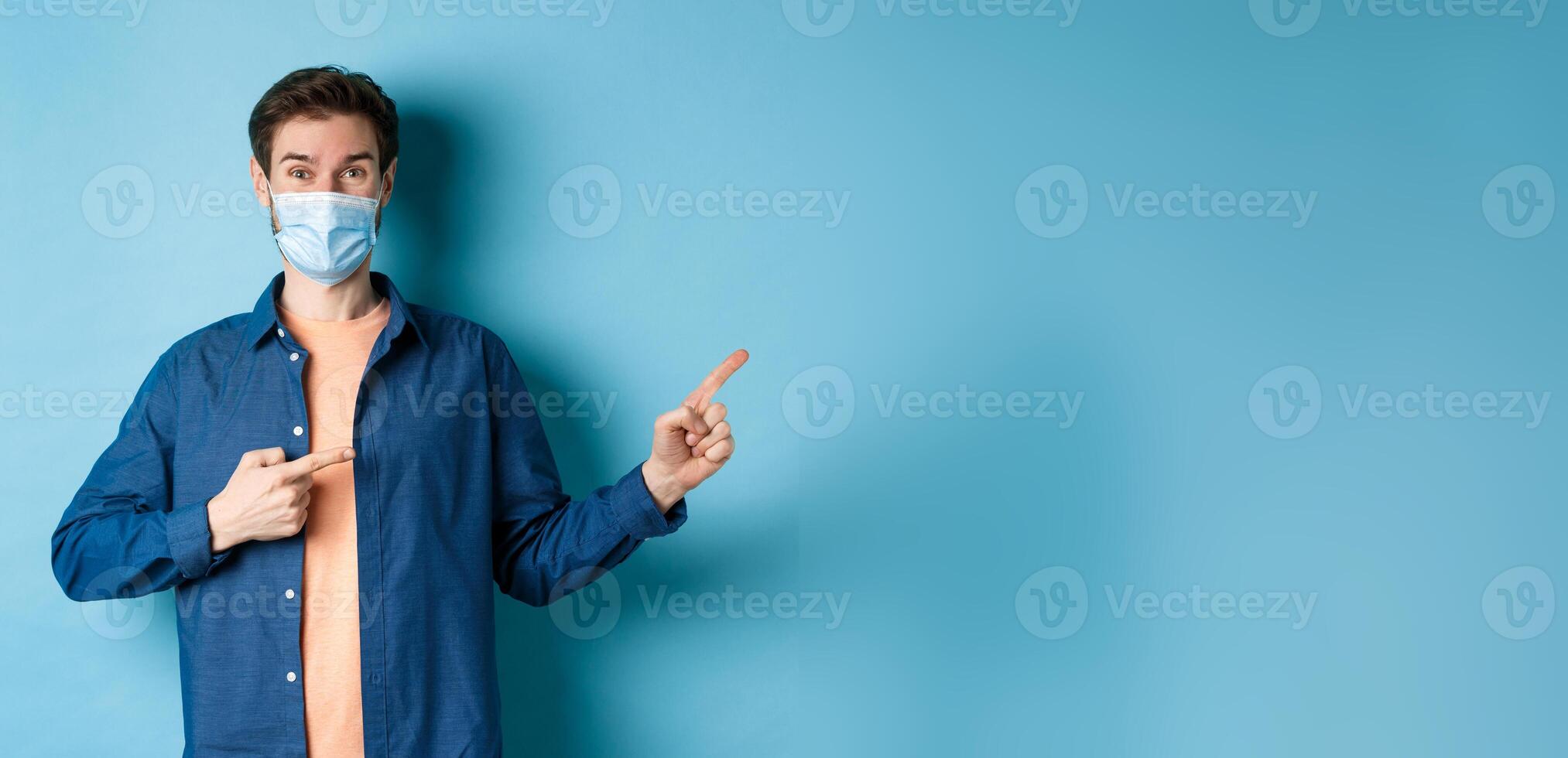 Covid-19 and pandemic concept. Happy healthy guy in medical mask pointing fingers at upper right corner, showing advertisement, standing on blue background photo