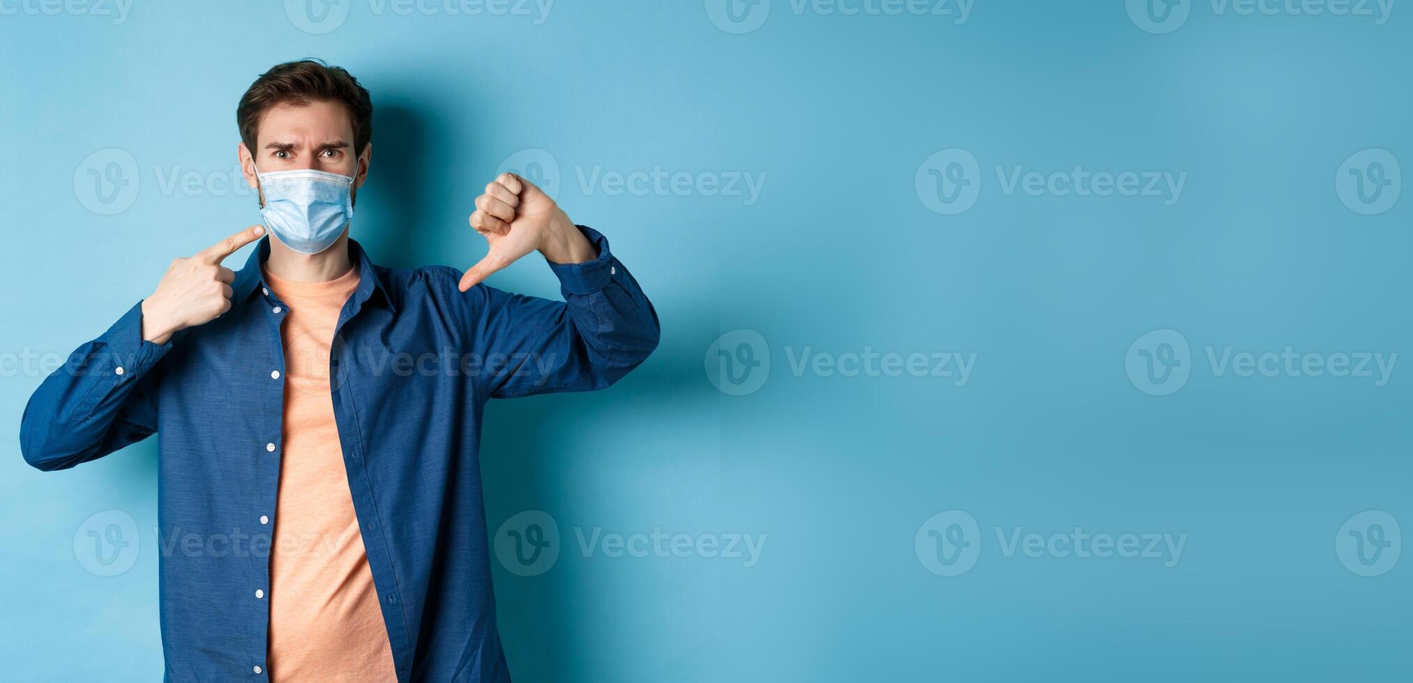 Coronavirus and pandemic concept. Disappointed young man pointing at medical mask and showing thumb down, disapprove and dislike quarantine rules, blue background photo