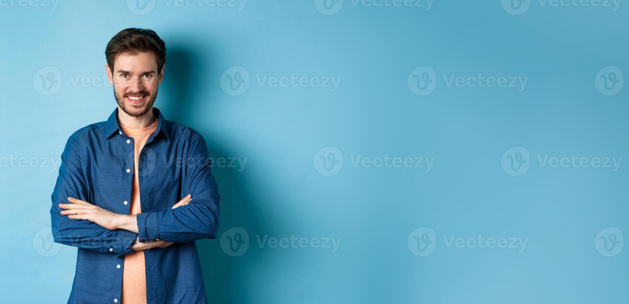 Confident young man feeling like professional, cross arms on chest and smiling sassy, standing on blue background photo