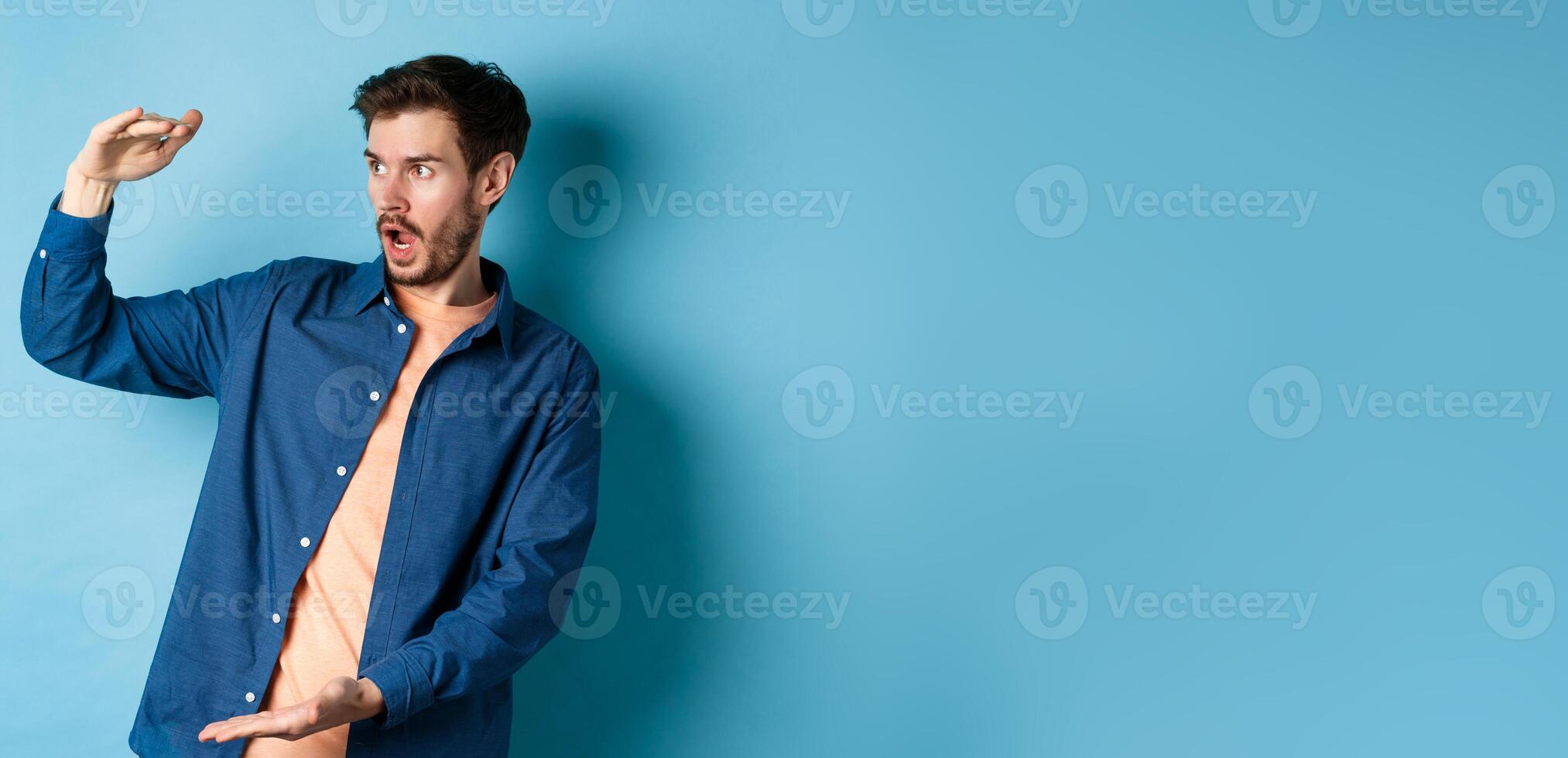Impressed caucasian guy staring at empty space with big object, shaping large size, standing on blue background photo