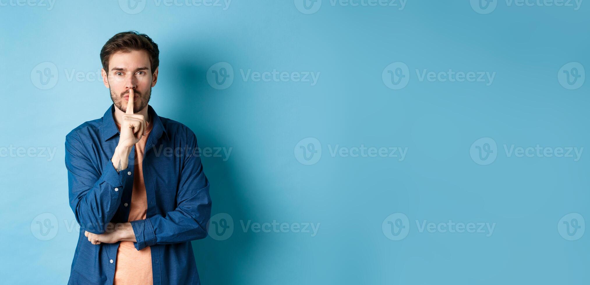 Modern guy shushing at camera, press finger to lips and telling a secret, making taboo sign, standing on blue background photo