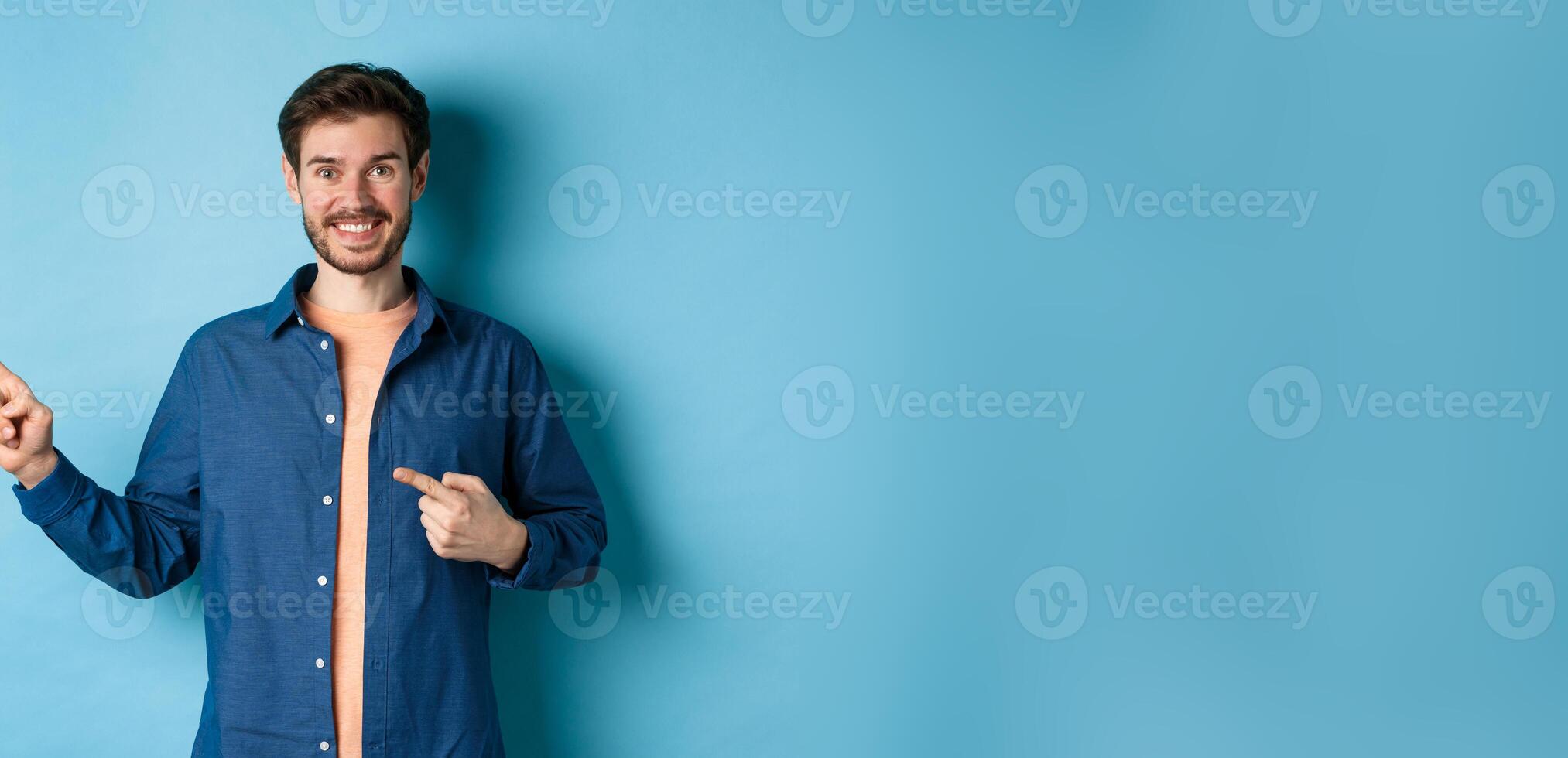 Happy smiling man pointing fingers left at logo, showing advertisement, standing on blue background photo