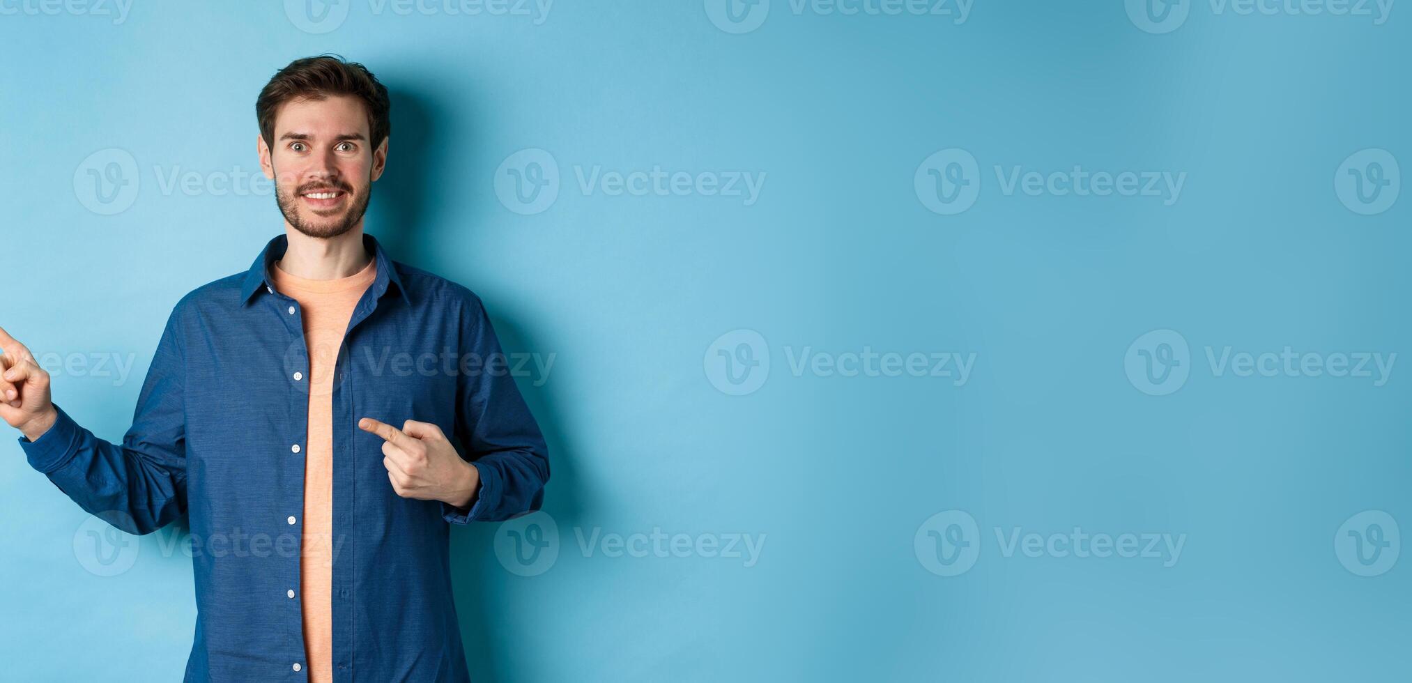 Happy smiling caucasian man showing advertisement, pointing fingers left at empty space, standing on blue background photo