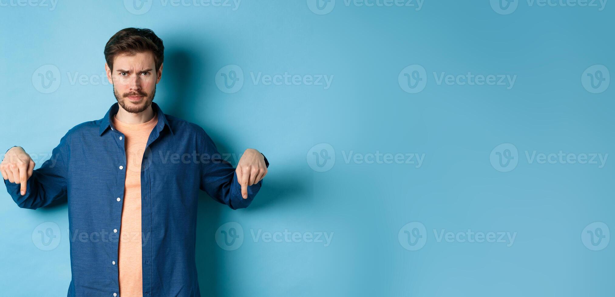 Concerned young man, frowning and looking worried, pointing fingers down, showing something bad, standing on blue background photo