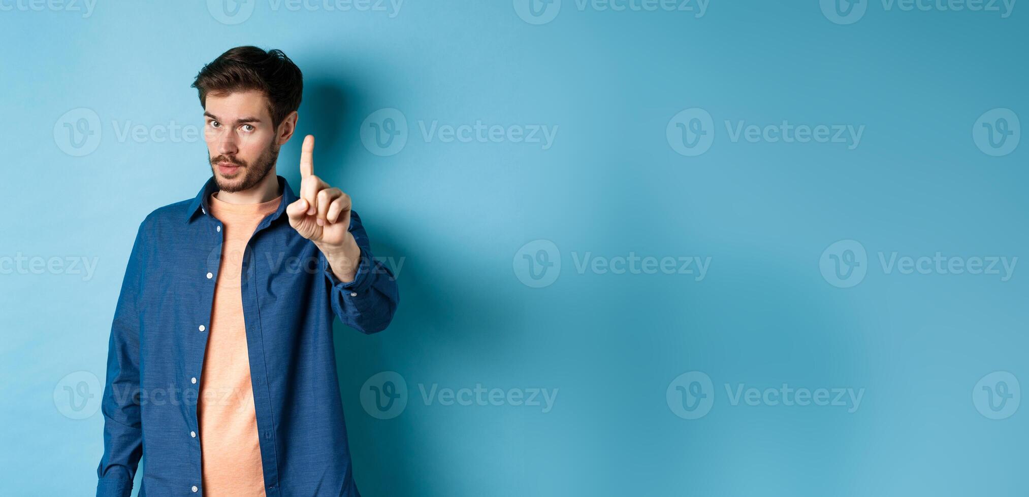 Image of young man give warning, teaching a lesson, raising one finger to scold, looking at camera, patronizing someone, standing on blue background photo