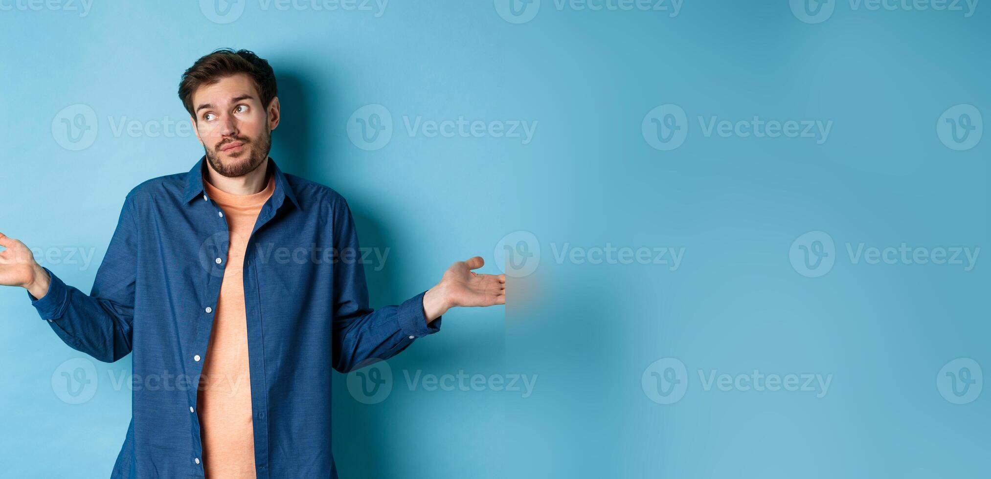 Indecisive young man with beard, look away and shrugging shoulders, know nothing, being clueless, standing on blue background photo