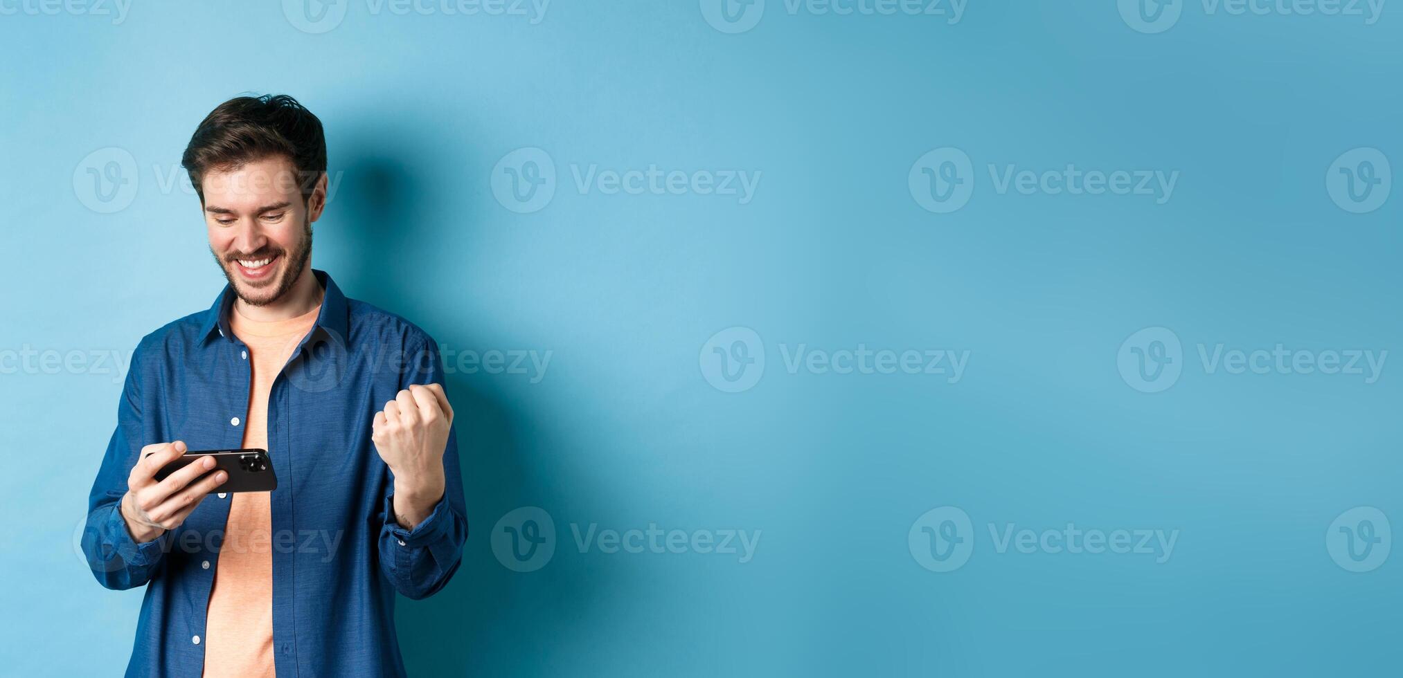Happy young man winning in game on mobile phone, looking satisfied at smartphone screen and say yes with joy, standing on blue background photo