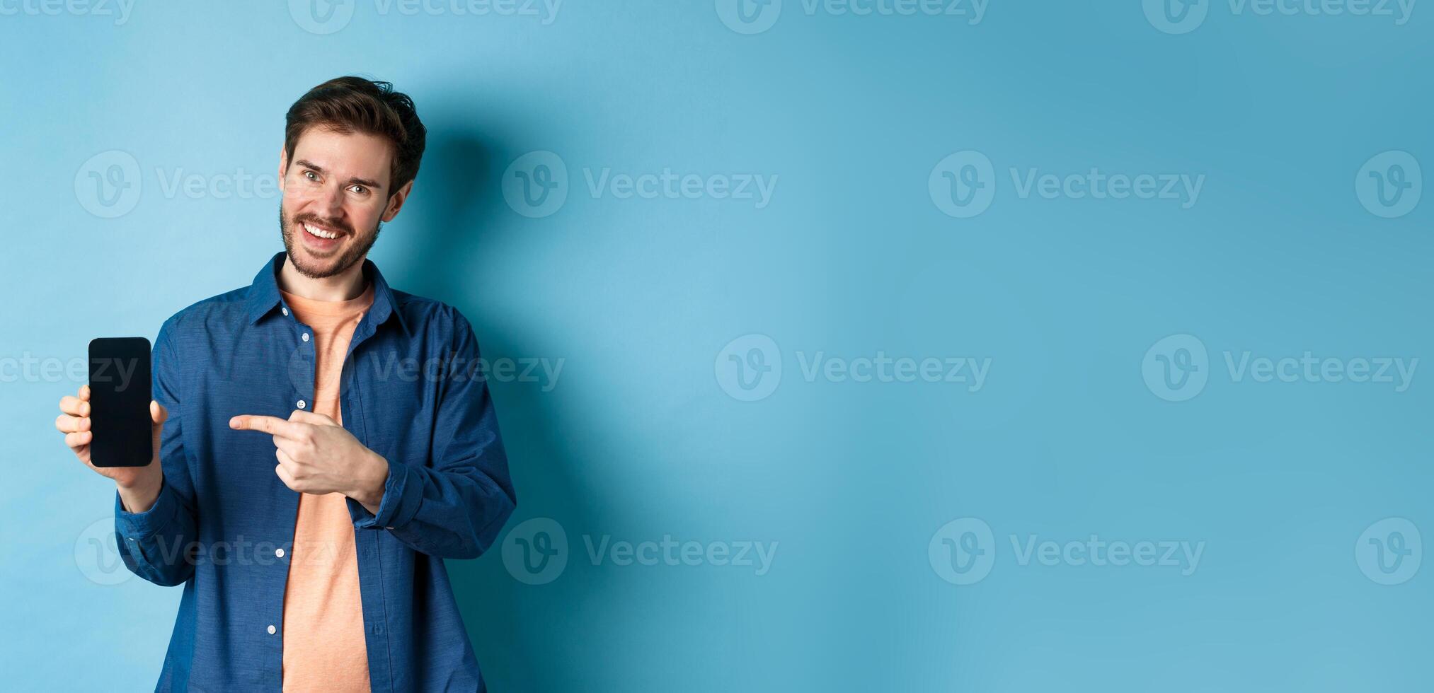 Smiling caucasian man showing on empty smartphone screen, pointing at mobile phone and looking satisfied, standing on blue background photo