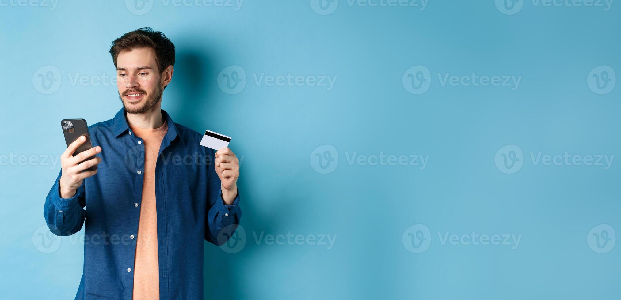 E-commerce concept. Image of handsome caucasian man paying online, looking at mobile phone screen and holding plastic credit card, blue background photo
