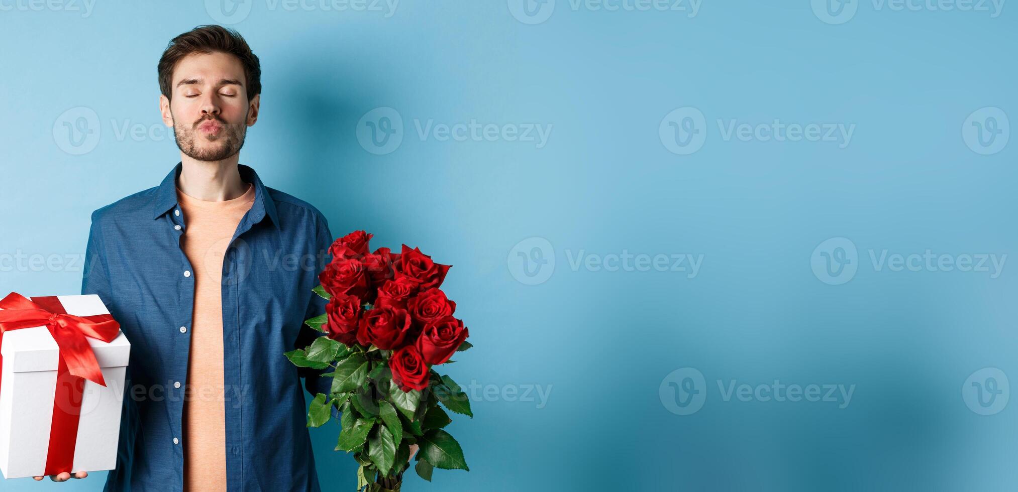 Love and Valentines day concept. Romantic man waiting for kiss, holding gift box and bouquet of red roses for lover on date, standing over blue background photo
