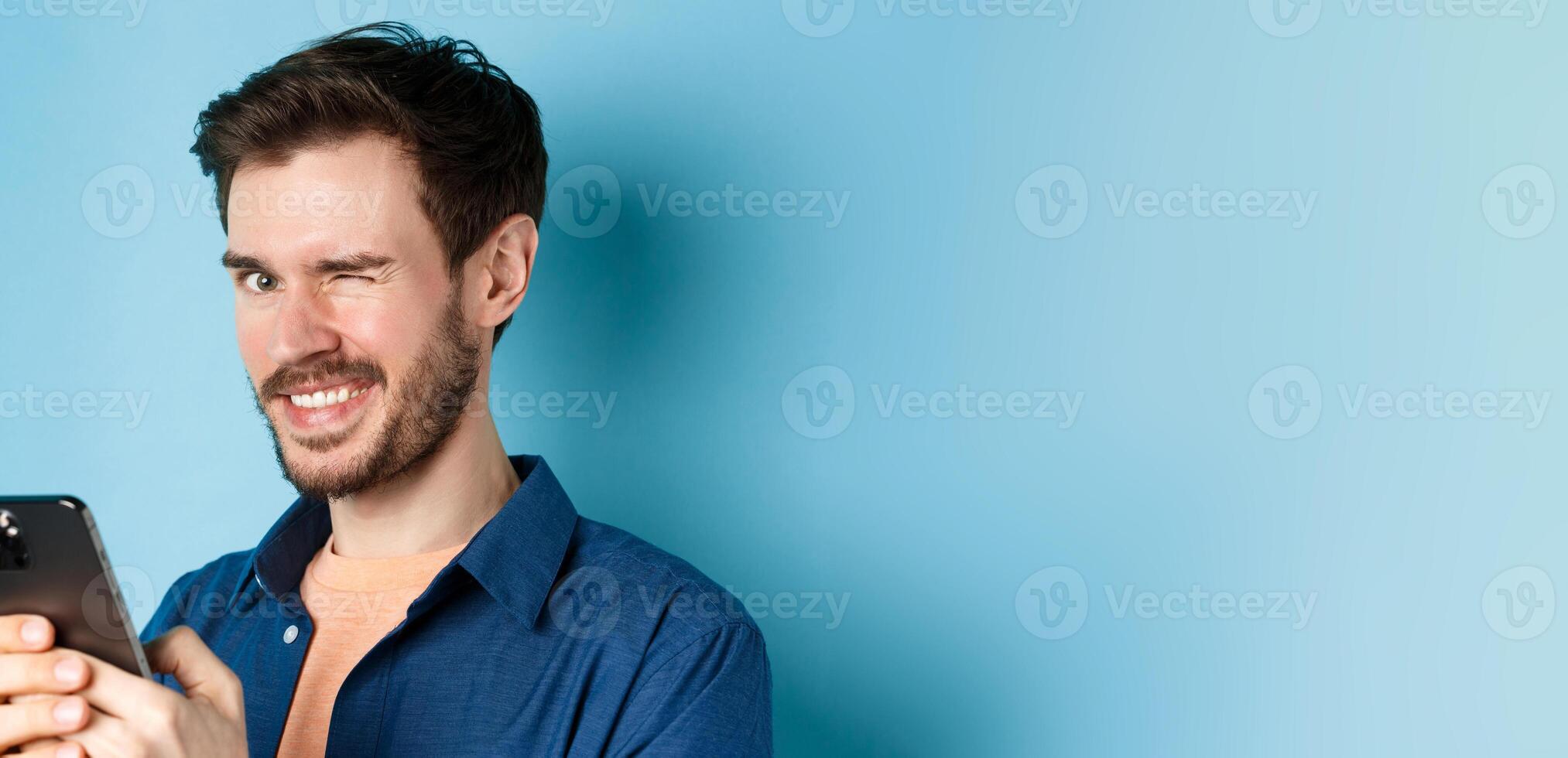 Portrait of handsome young man with beard, winking and smiling at camera, using smartphone, standing on blue background photo