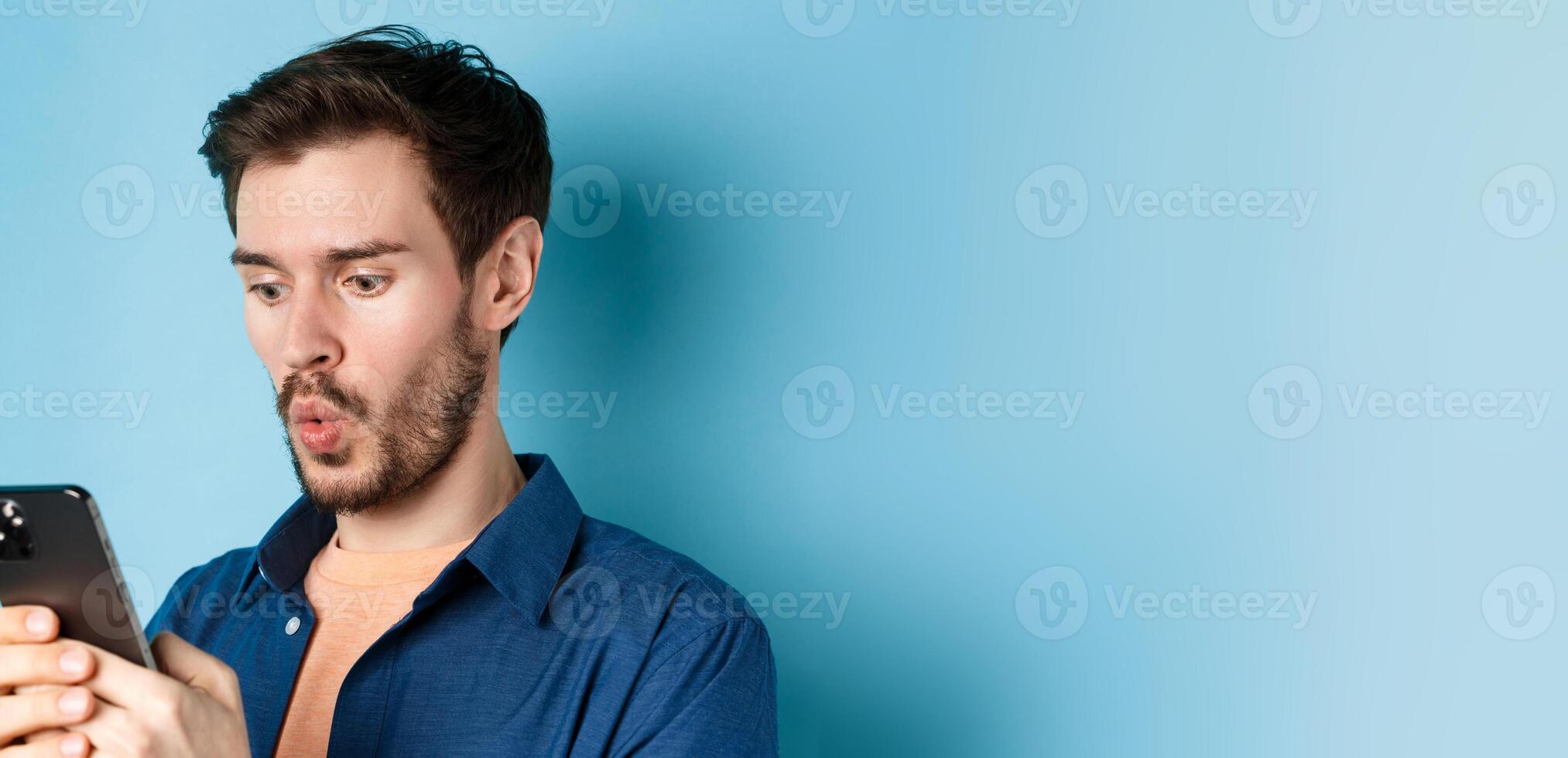 Image of impessed young man looking at smartphone and saying wow, checking out online offer, standing on blue background photo