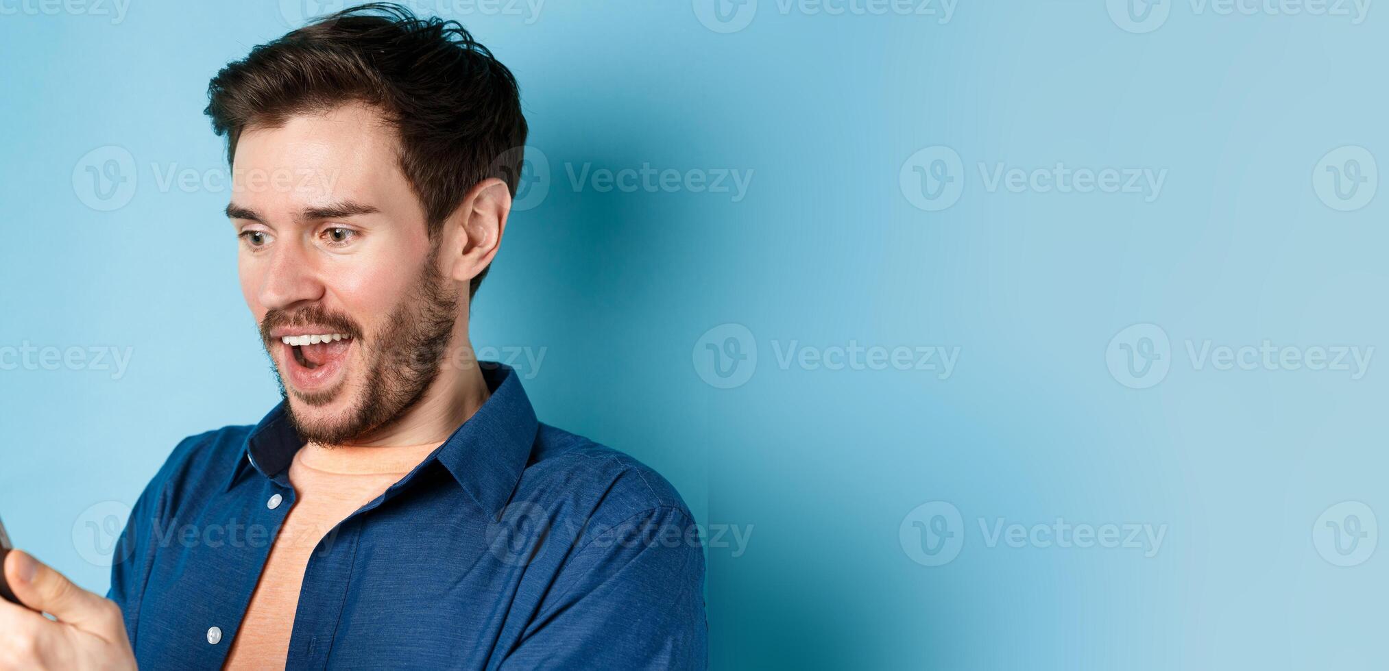 Image of handsome young man feeling happy reading good news on mobile phone, standing cheerful against blue background photo