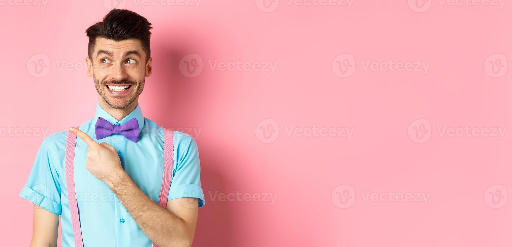 Happy smiling man in funny outfit and bow-tie, pointing finger left and looking at logo, showing advertisement on pink background photo