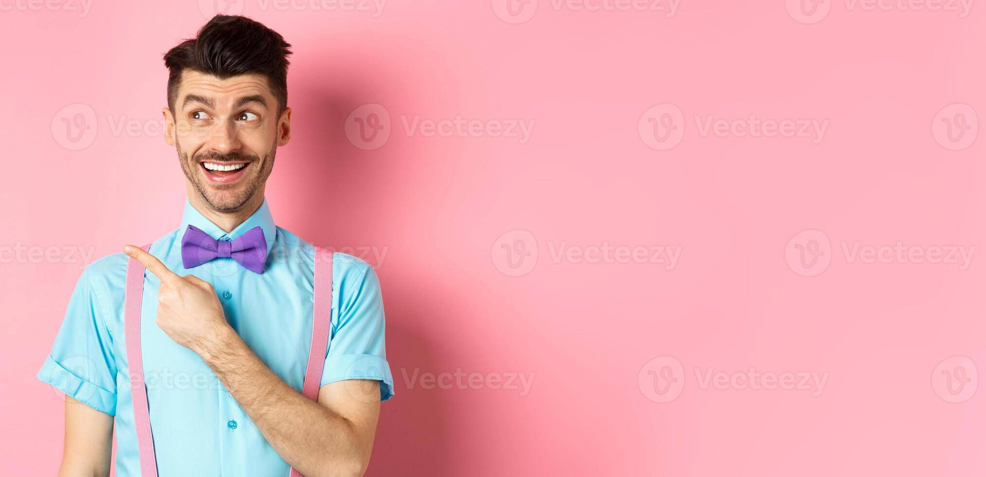 Cheerful caucasian man in funny bowtie looking and pointing hand left, showing promo logo, standing on pink background photo