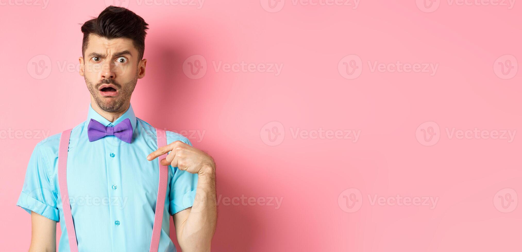 Image of confused funny guy in bow-tie and suspenders, pointing at himself as if being accused or chosen, raising eyebrow surprised, standing over pink background photo