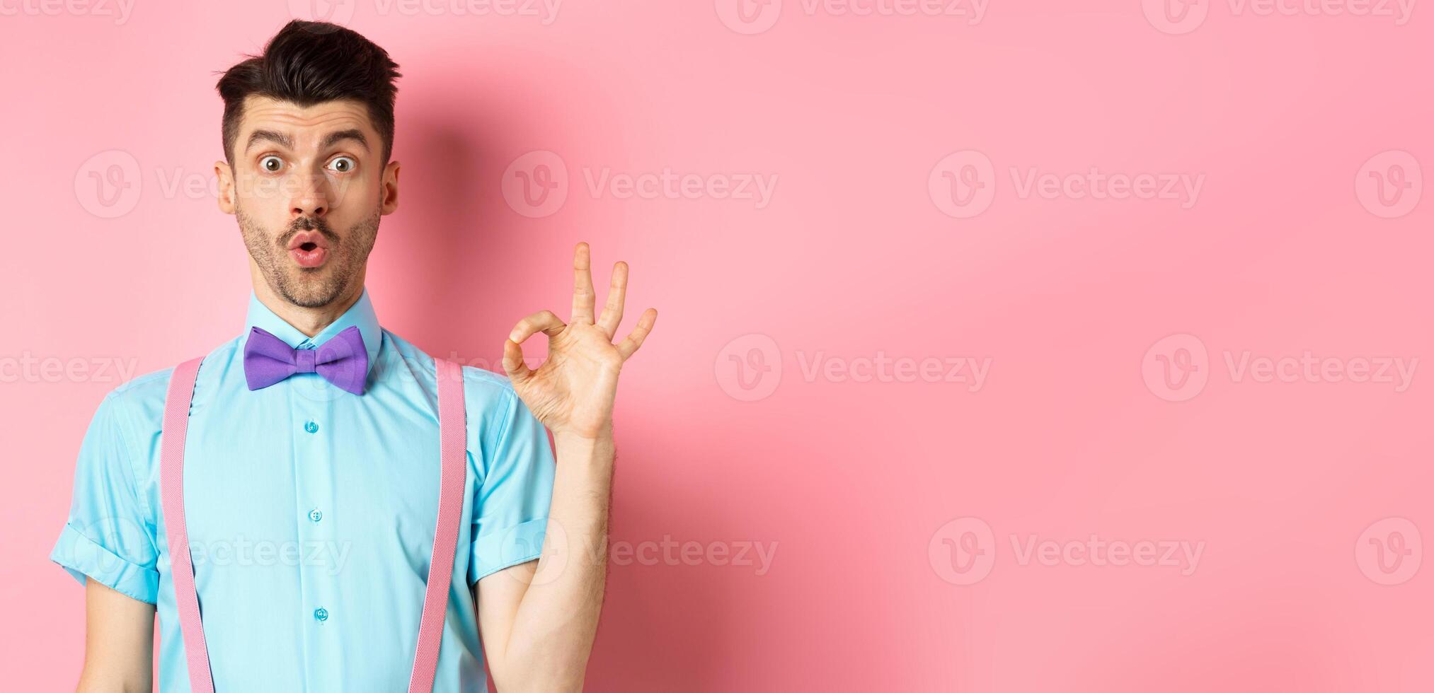 Impressed guy in bow-tie and suspenders saying wow, showing okay gesture with amazement, praise awesome thing, standing over pink background photo