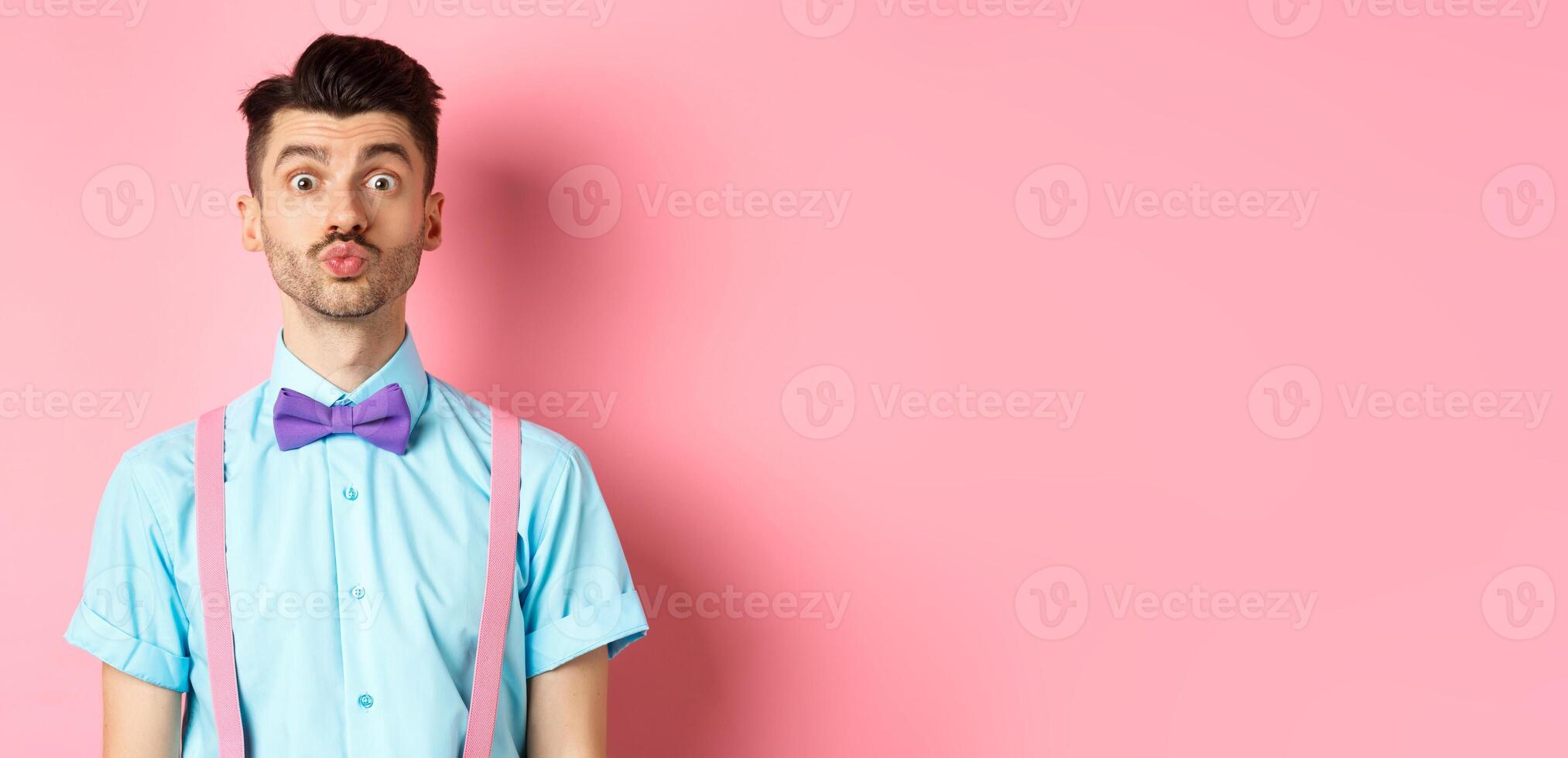 Funny young man waiting for kiss with puckered lips and silly face, standing on romantic pink background. Concept of love and Valentines day photo
