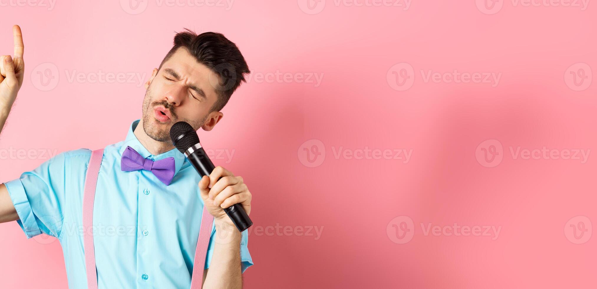 Party and festive events concept. Funny guy singing in microphone, raising finger up as reaching high note in song, standing on pink background photo