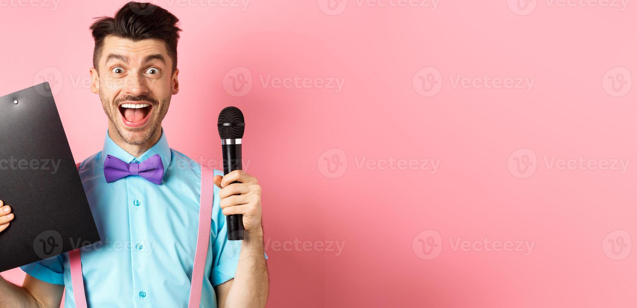 Holidays concept. Happy young man entertainer smiling at camera, holding clipboard and microphone, making speech on party event, standing over pink background photo