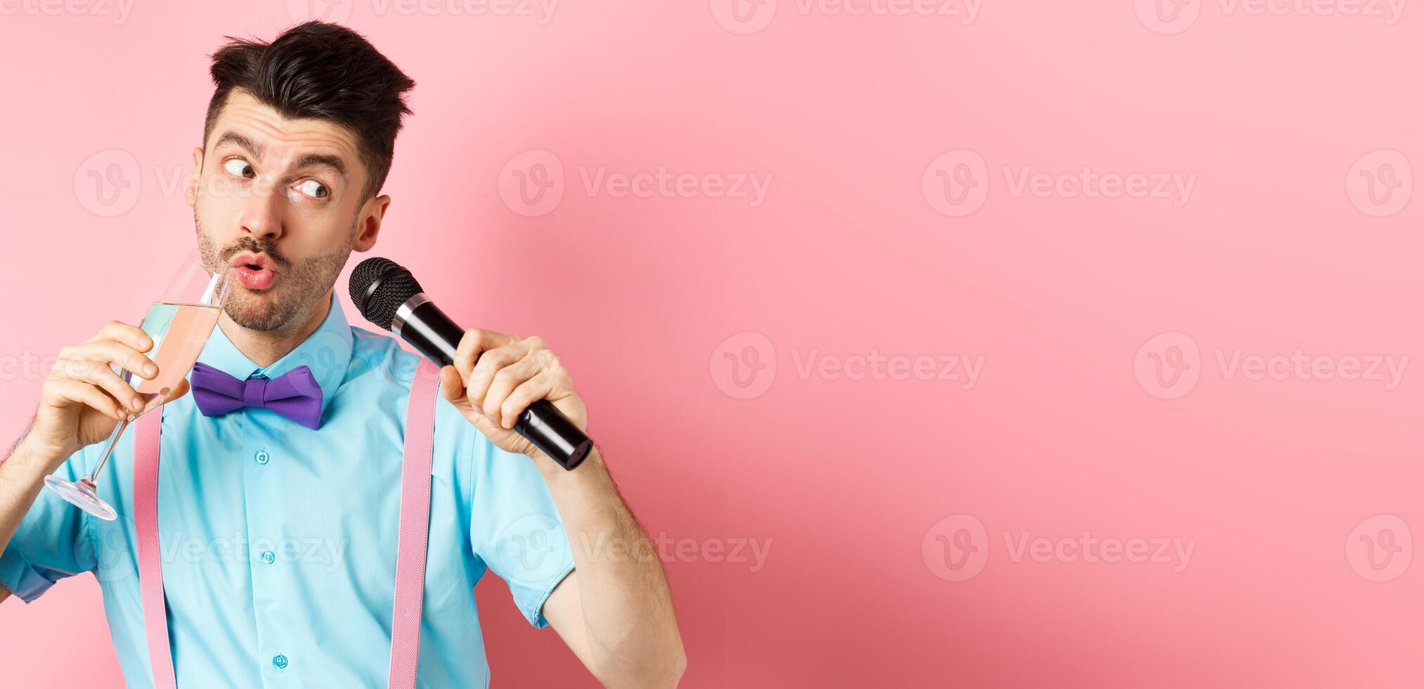 Party and festive events concept. Funny guy singing karaoke, performing song with microphone and drinking champagne from glass, standing on pink background photo
