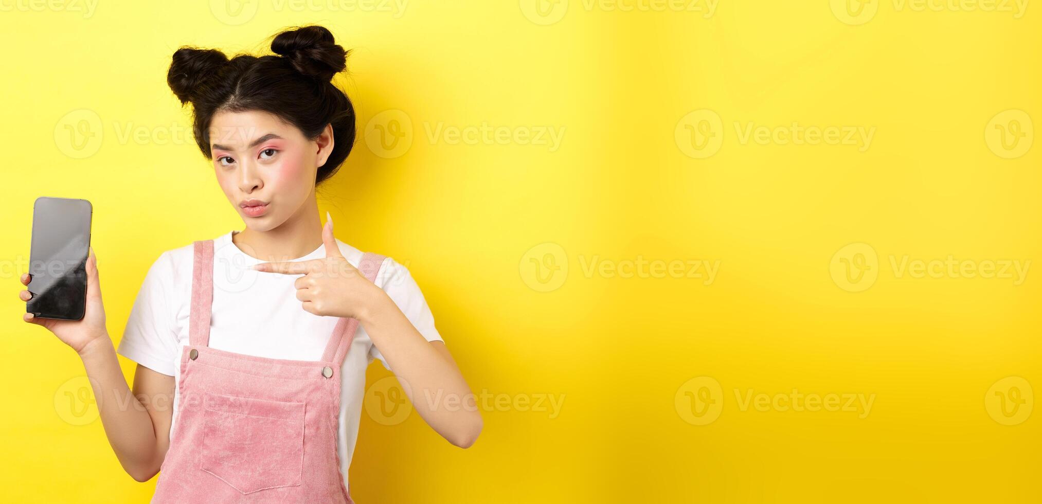 Online shopping concept. Stylish asian woman with pink makeup, pointing at empty smartphone sreen, showing good deal, yellow background photo