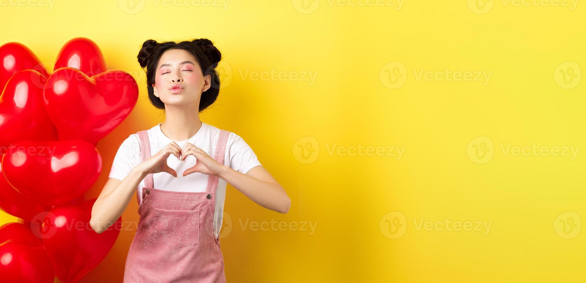 Romantic girl close eyes and pucker lips for kiss, showing I love you heart gesture, standing near cute red balloons, kissing lover over yellow background photo